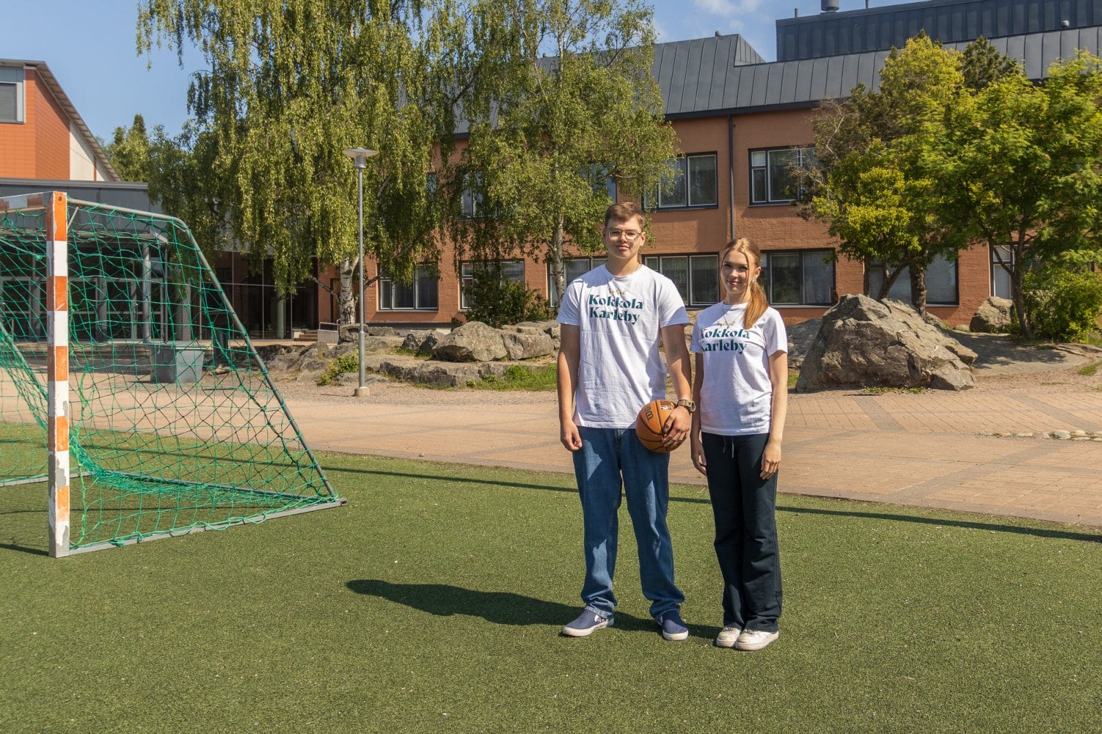 Nuori poika ja tyttö seisovat koripallokentällä, pojalla kädessään koripallo.