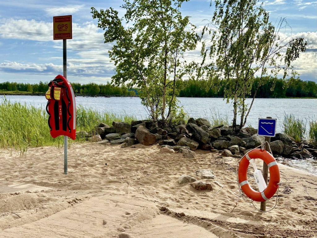 Potinrannan uimarannalla kirkkaanoranssi pelastuslautta sekä pelastusrengas hiekalla, taustalla vihreitä koivuja ja kivikkoa merta vasten.