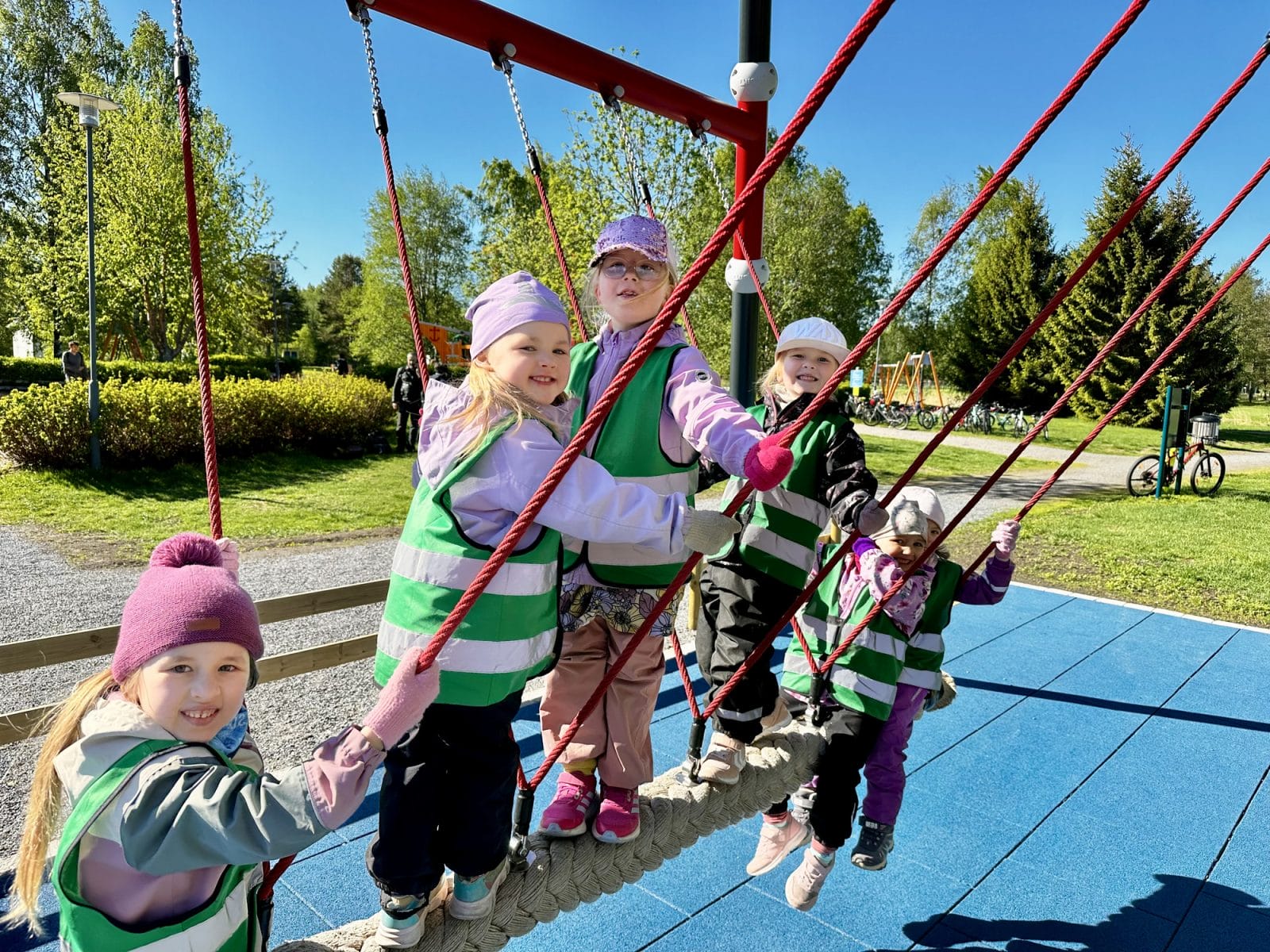 Ryhmä päiväkotilaisia istuu ja seisoo tukkilaiskeinun kyydissä aurinkoisessa puistossa.