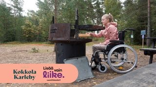 Henkilö istuu nuotiopaikan äärellä pyörätuolissa ja laittaa halkoja nuotioon.