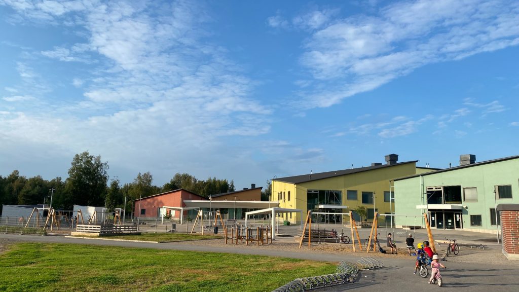 Isokylän koulurakennus ja sen piha-alue kesäpäivänä. Pihalla lapsia pyöräilemässä. Kaksi lasta istuu keinuissa.