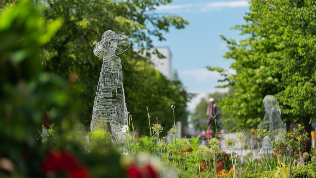 I förgrunden en ståltrådsskulptur som föreställer en kvinna med hatt med brätte. Omkring finns blommor och träd.