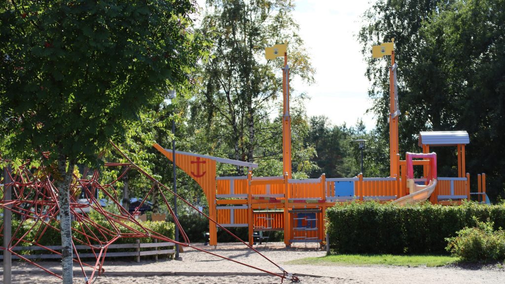Lekplats för barn i sanden i Havsparken, omgiven av träd. I mitten på bilden syns en klätterställning i form av ett fartyg med sina rutschbanor. Till vänster, i bildens förgrund, syns en klätterställning av rep.