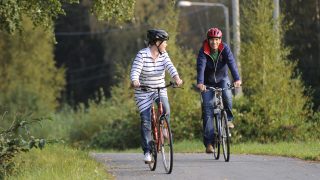 En man och en kvinna cyklar längs en cykelväg. Kvinnan, som cyklar lite före, är iklädd en randig tunika, jeans och vita tennistossor. Mannen, som kommer efter, är iklädd jeans och en mörkblå munkjacka som är delvis öppen. Båda har en cykelhjälm på sig. Kvinnan tittar leende mot mannen. I bakgrunden syns lummiga lövträd.