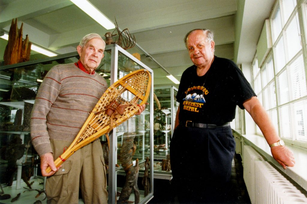 Salkio holds a wooden snowshoe in his hand. Next to him Ray Rinta is resting on window sill.