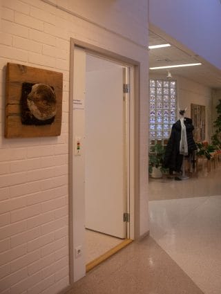 Sisätila, jossa valkoinen tiiliseinä. Kuvassa keskellä valkoinen ovi auki toimistotilaan.