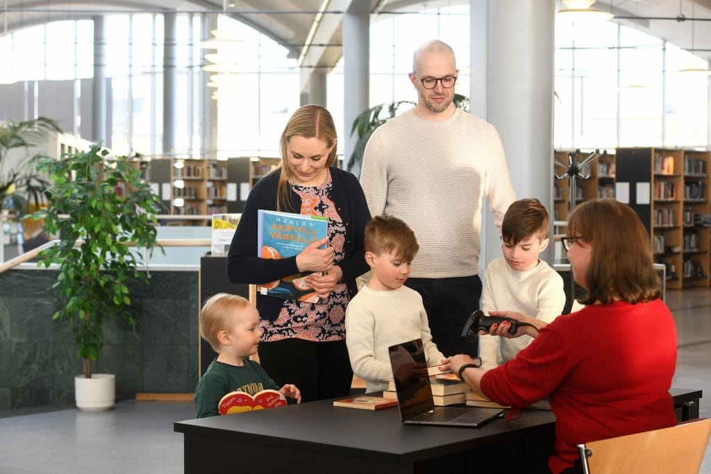 Två vuxna och tre barn lånar böcker vid biblioteket. En bibliotekarie sitter bakom disken och lånar ut böcker.