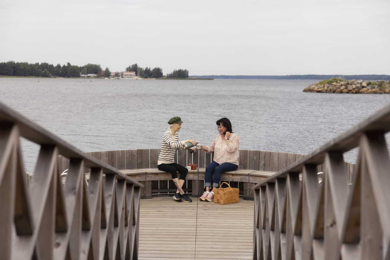 Kaksi naista istuu laiturilla eväskorin kanssa. Toinen kaataa kahvia toisen kädessä olevaan kuppiin. Taustalla näkyy meri ja aallonmurtaja.