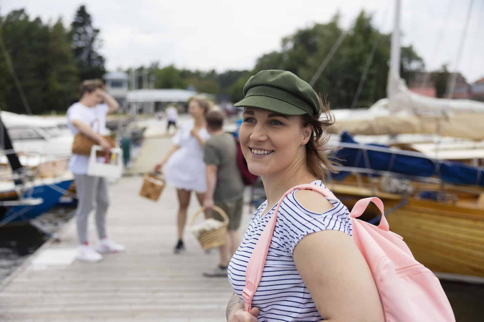 Vihreään lakkiin pukeutunut nuori nainen seisoo laiturilla vaaleanpunainen reppu olallaan. Taustalla näkyy veneitä ja ihmisiä eväskorit kädessään.