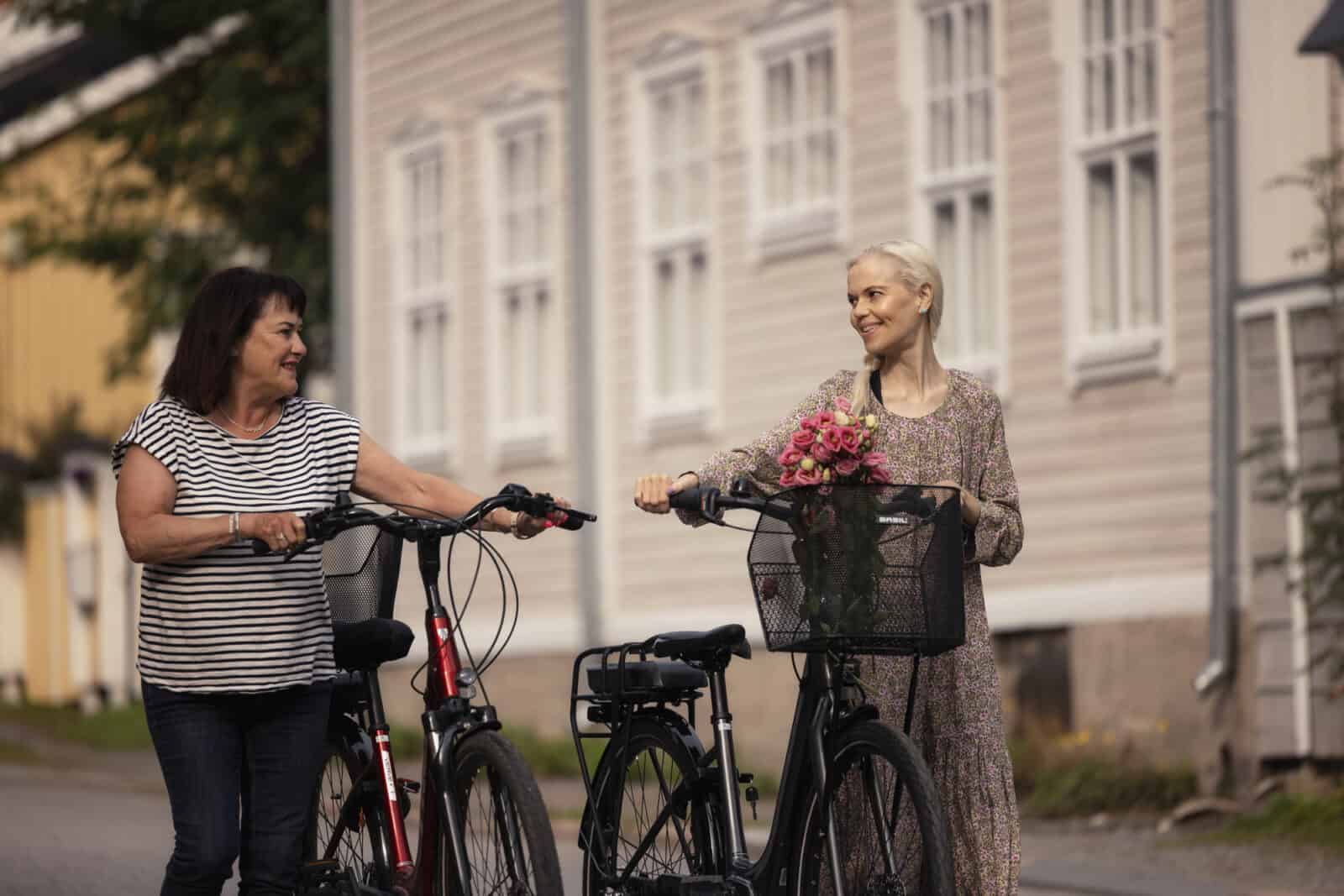Kaksi naista taluttaa pyöriä. Toisen pyörän korissa on kukkia. Naiset hymyilevät, taustalla näkyy vaaleanpunainen puutalo.