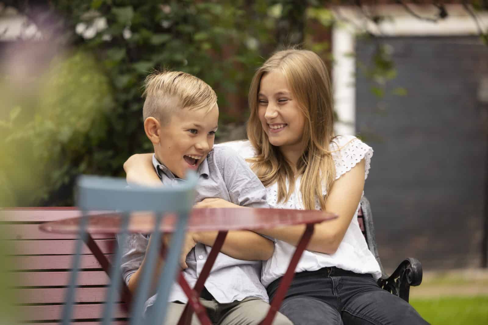 Tyttö ja poika istuvat puutarhassa. He nauravat yhdessä. Etualalla näkyy puutarhatuoli, takana piharakennuksen ovi.