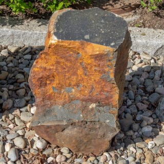 Kiepin kivilajinäyttelyn näytelohkare. Tumma kivi, jonka pinnoista osa ruostunut.