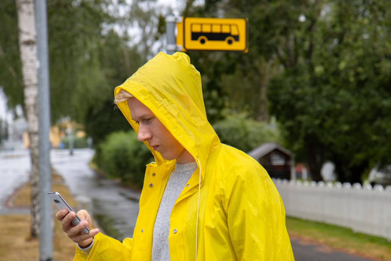 Poika keltaisessa sadetakissa odottaa bussia pysäkillä ja selailee kännykkää